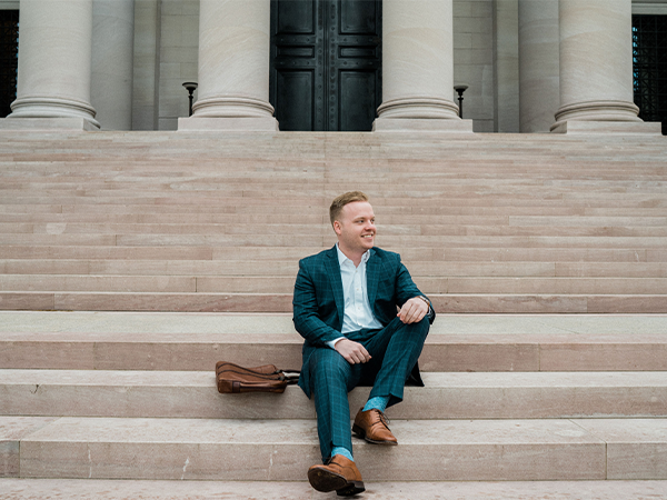 A person in a suit sits on the marble steps of a stately building, his leather bag sitting beside him.